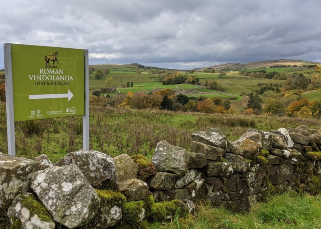 The Roman fort of Vindolanda pre-dates Hadrian. In this picture it is in the centre, level with the signpost's arrow. Hadrian's Wall is on the horizon, right.